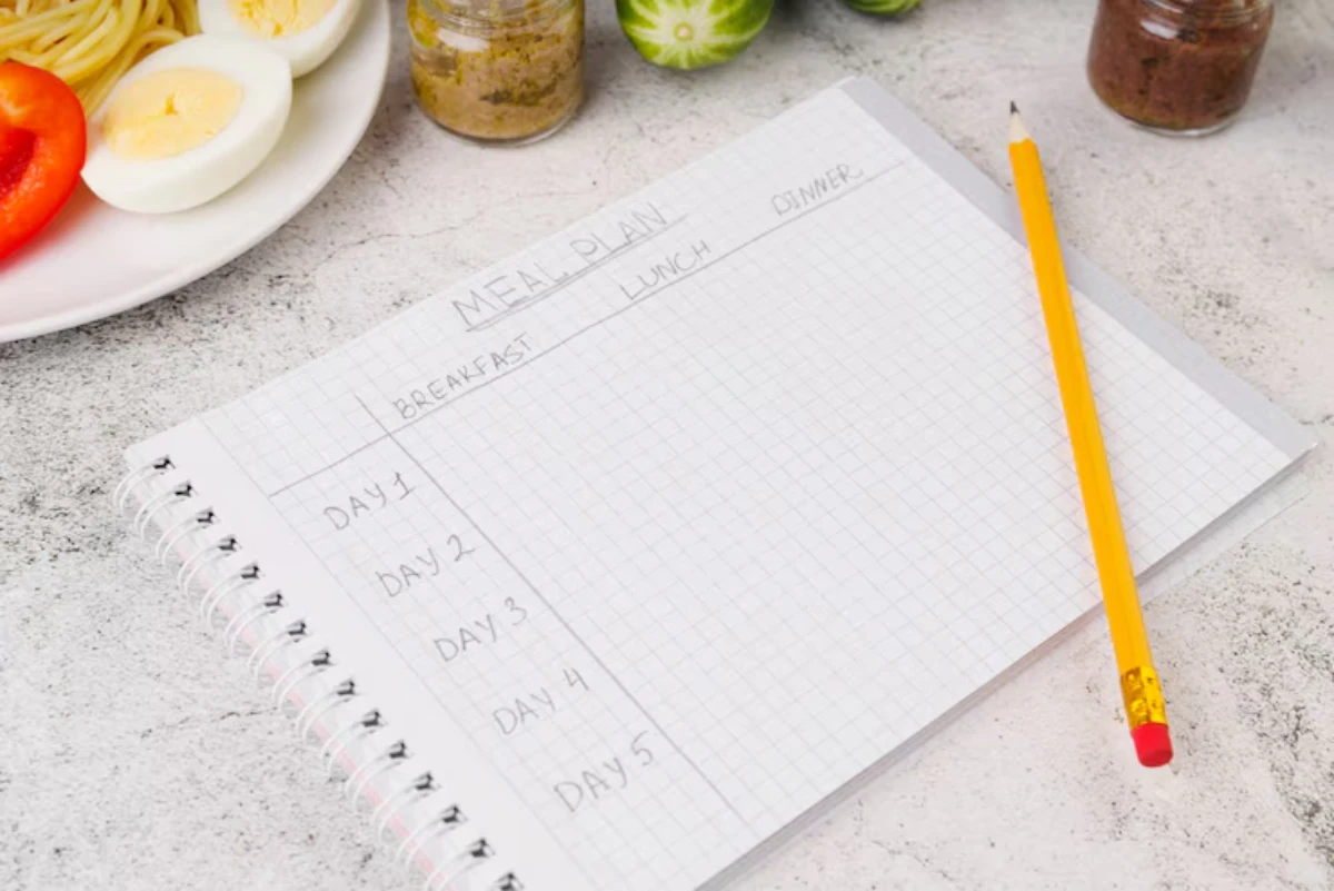 A meal planning notebook with a grid layout, a yellow pencil, and a plate of pasta, boiled eggs, and vegetables in the background.