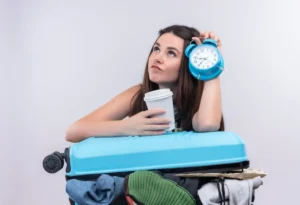 A person holds a blue alarm clock in one hand and a coffee cup in the other, leaning on a suitcase filled with clothes.