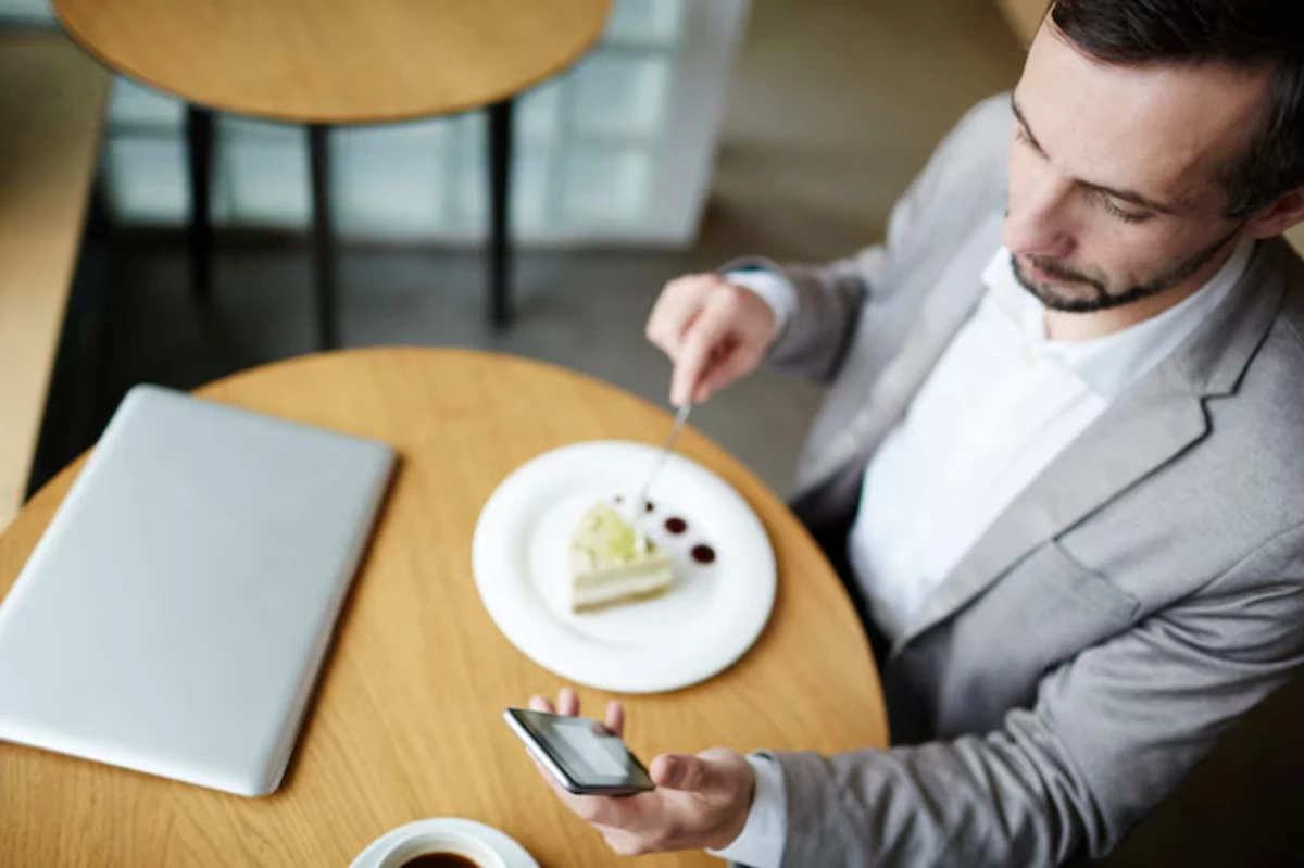 A person in a suit sits at a wooden table, enjoying cake and coffee while using a smartphone alongside a laptop.