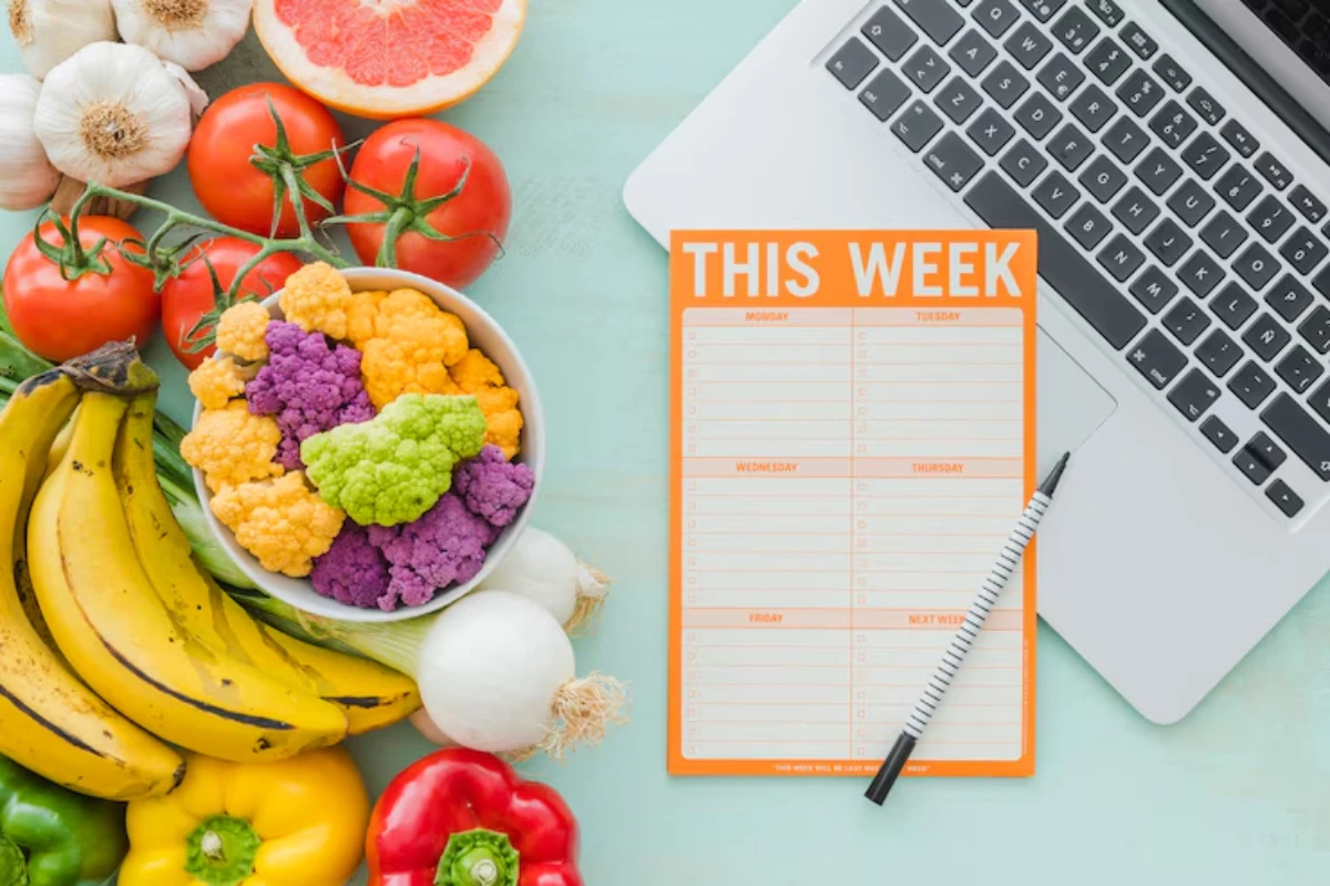 A colorful array of fresh vegetables and fruits surrounds a weekly planner and a laptop, emphasizing healthy meal planning.