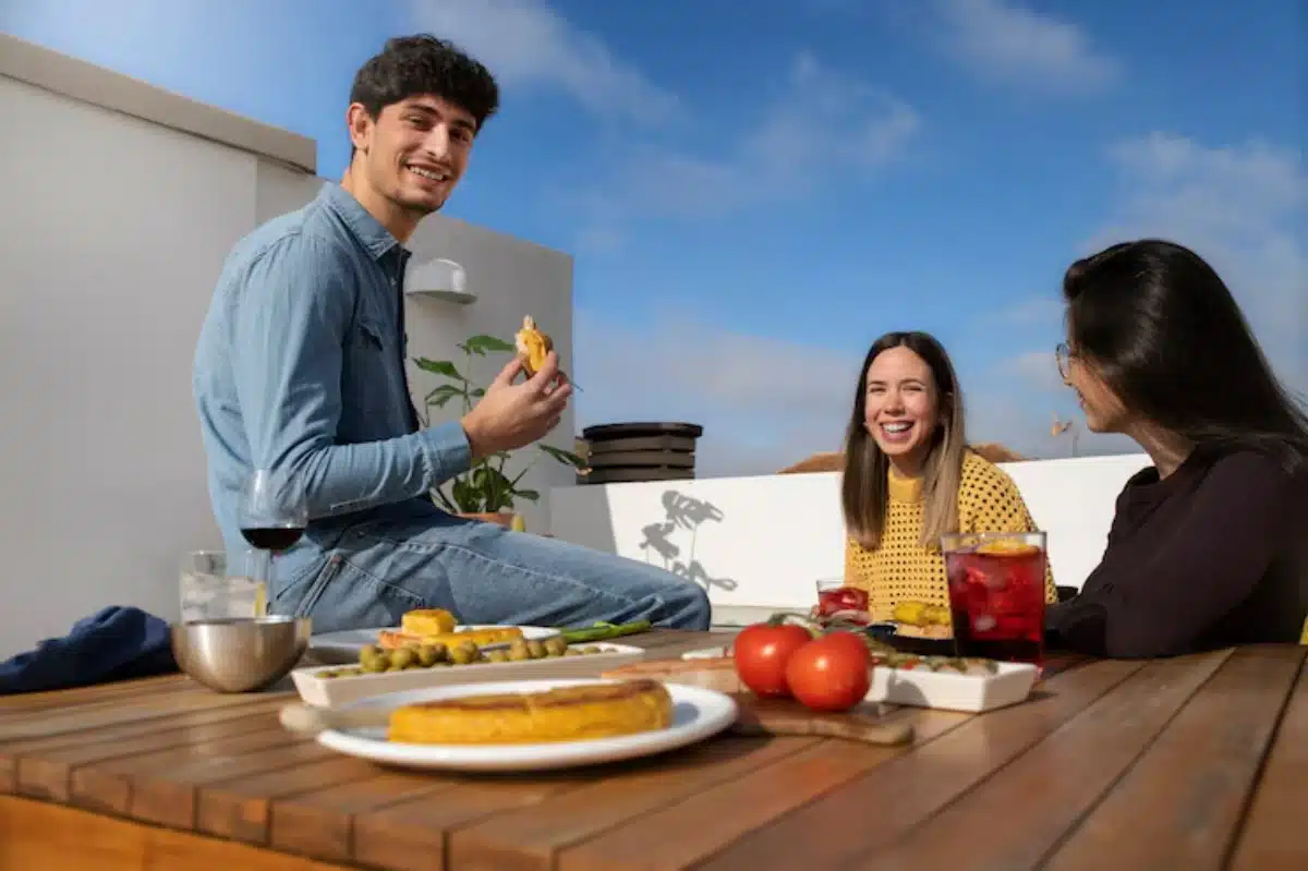 A outdoor gathering featuring a table with food, drinks, and three people enjoying a sunny day against a blue sky.