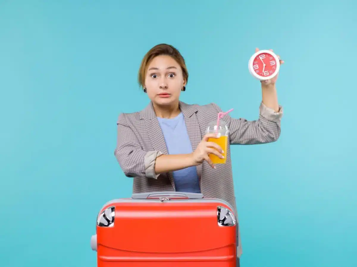 A person holds a red clock and a glass of orange drink near a bright red suitcase against a blue background.