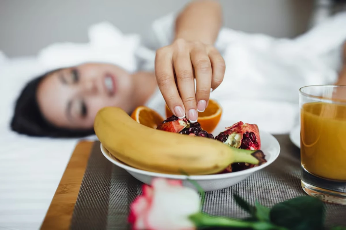 A woman reaches for fresh fruit, including a banana and pomegranate, on a tray beside orange juice and a rose while lying in bed.