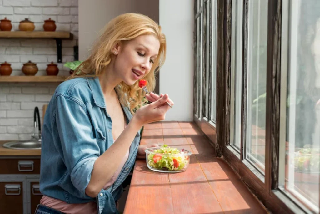 A woman with wavy blonde hair enjoys a fresh salad while seated at a wooden table next to large windows.