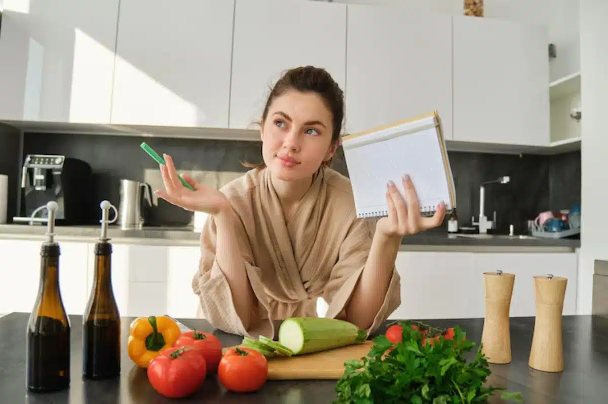 A person in a kitchen, wearing a robe, holds a notebook in one hand and gestures with a pen, surrounded by fresh vegetables and bottles.