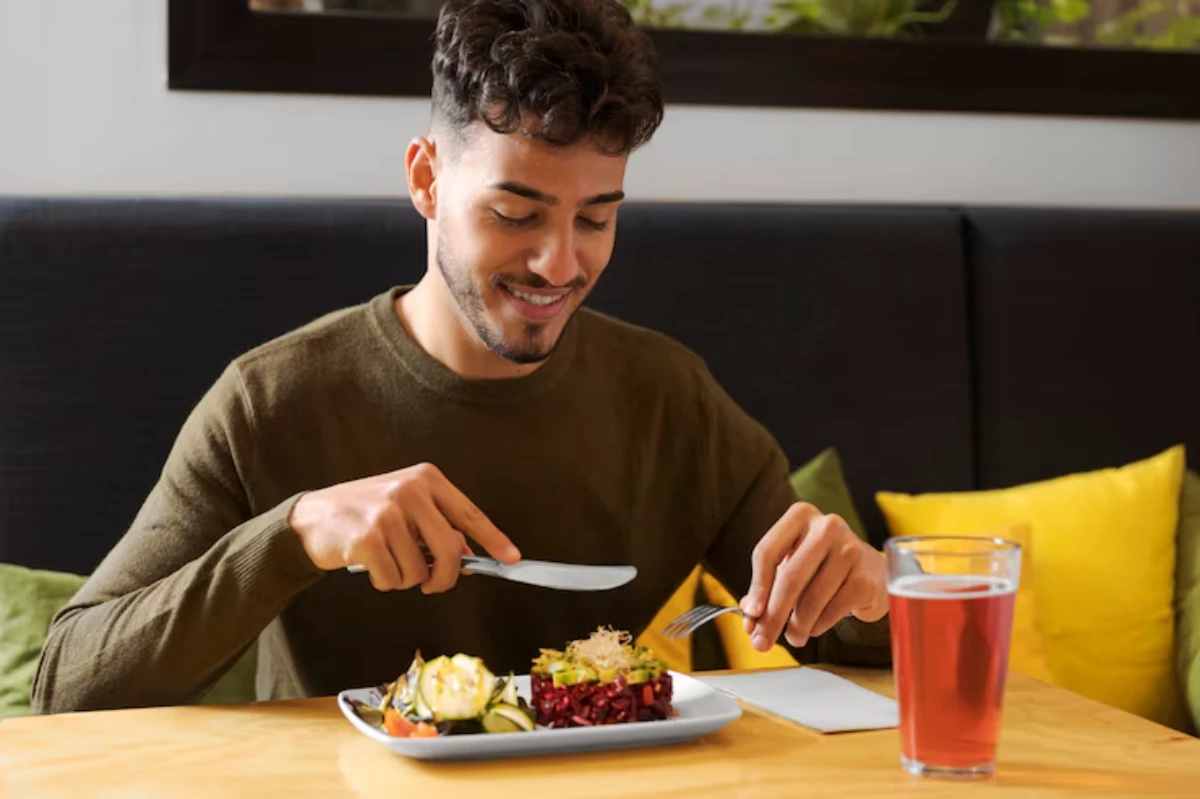A person sits at a wooden table, preparing to eat a colorful plate of salad and vegetables, with a glass of pink drink nearby.