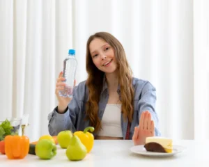 A person holds a water bottle with one hand while gesturing to refuse a slice of cake on a table filled with fresh fruits and vegetables.