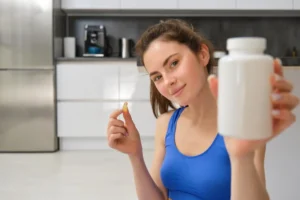 A person in a blue sports bra holds a vitamin capsule in one hand and a white supplement bottle in the other, seated indoors.