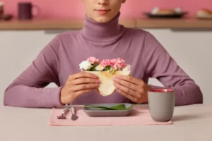 A person in a lavender turtleneck holds a sandwich with flowers, seated at a table with a gray dish, fork, and mug against a pastel backdrop.