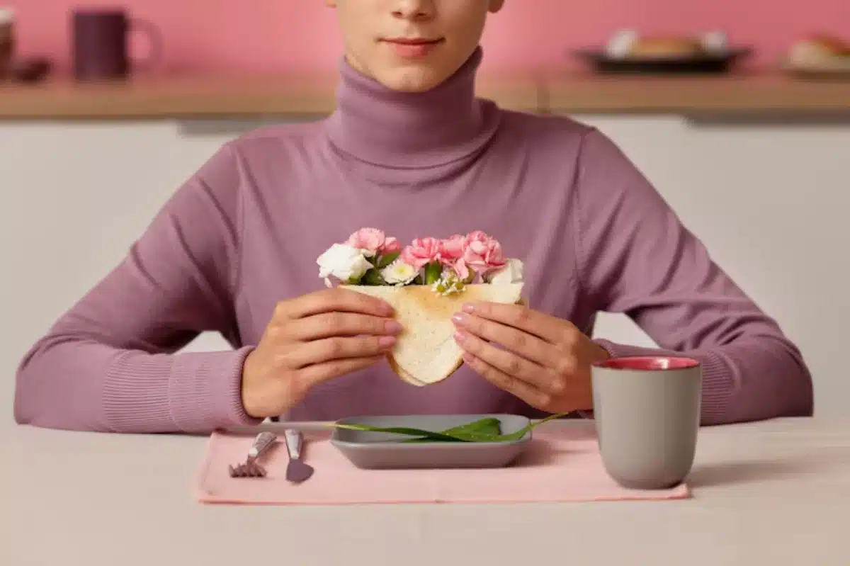 A person in a lavender turtleneck holds a sandwich with flowers, seated at a table with a gray dish, fork, and mug against a pastel backdrop.