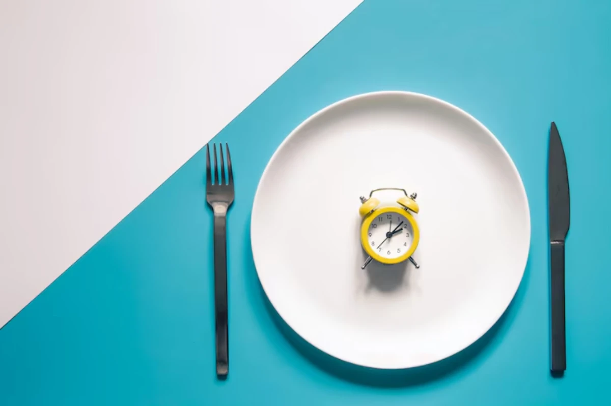 A white plate with a yellow alarm clock in the center, flanked by a black fork on the left and a knife on the right, on a teal background.