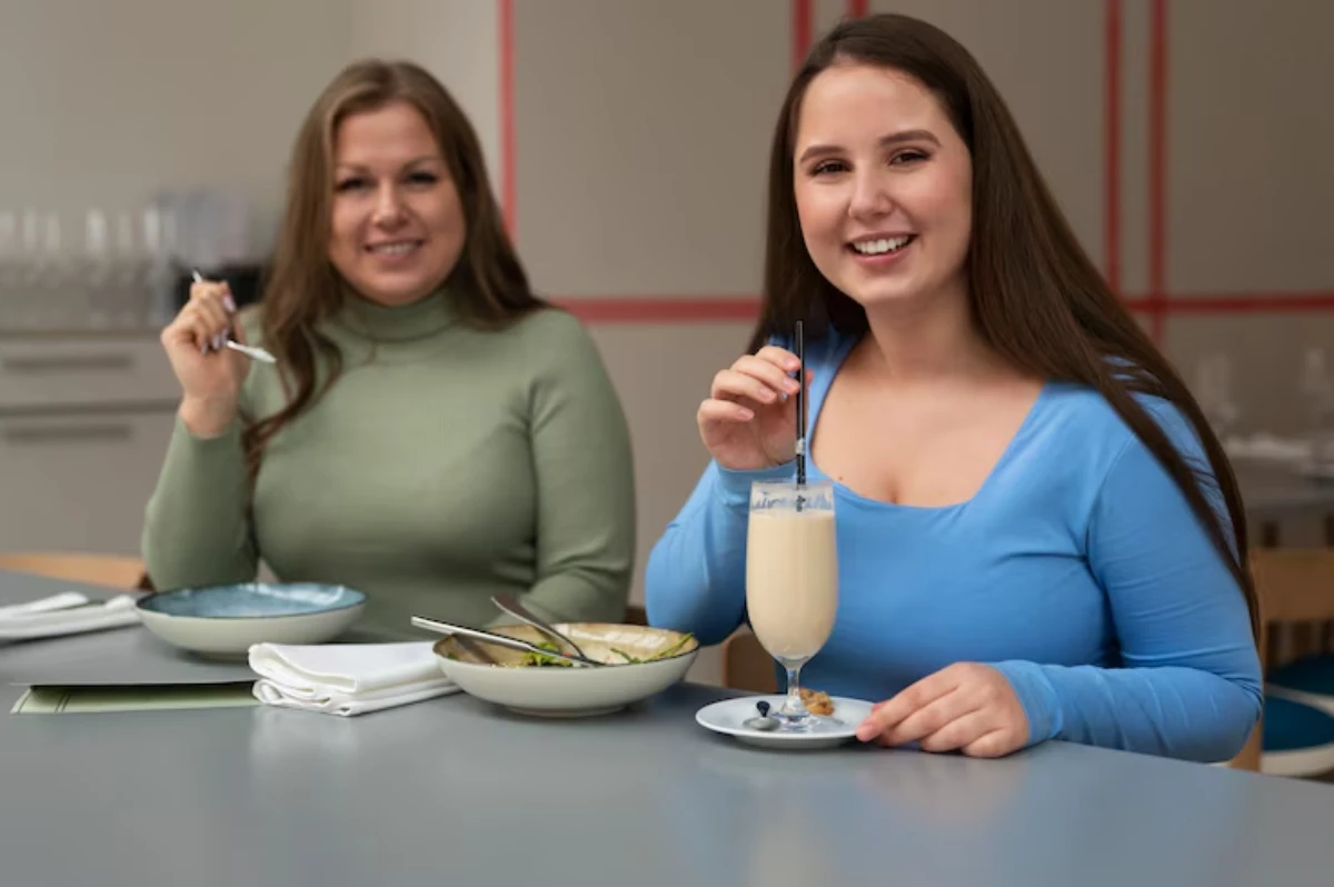 Two women sit at a table, enjoying a meal. One holds a drink with a straw, while the other has a spoon, surrounded by dishes.