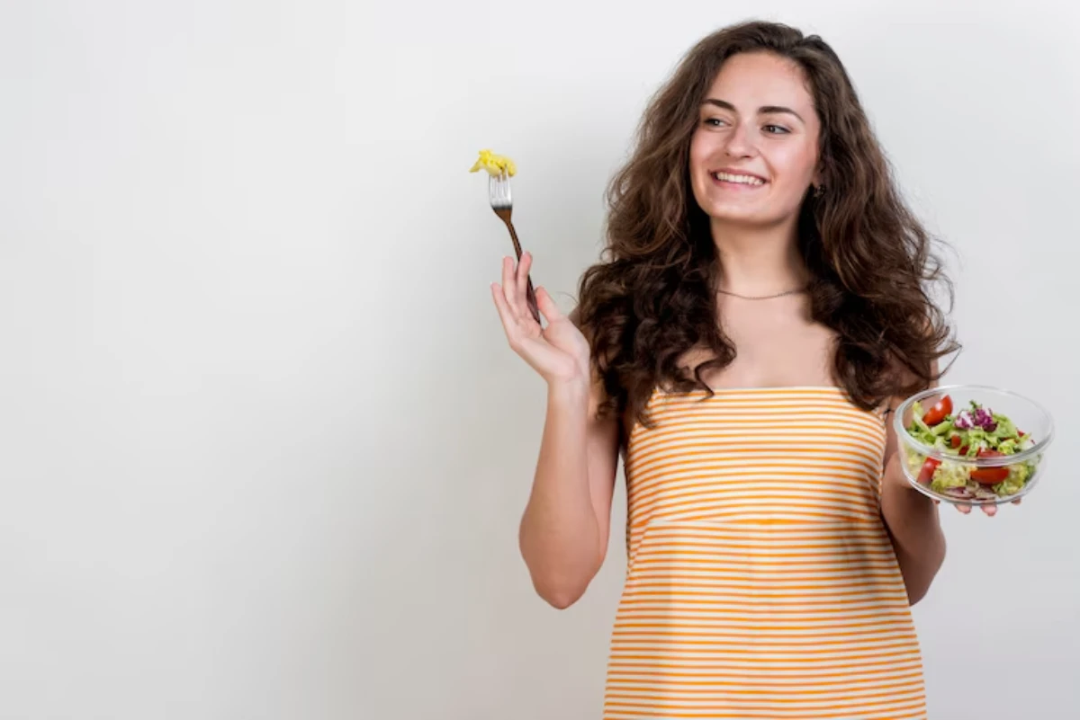 A person with curly hair holds a fork with pasta in one hand and a bowl of salad in the other, standing against a white background.
