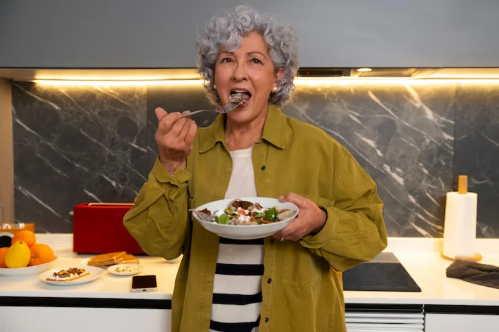 An elderly person in a green jacket holds a plate of food in a modern kitchen, ready to take a bite. Bright countertop decor is visible.