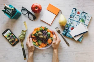 Aerial view of a fresh salad in hands, surrounded by stationery, an apple, a pear, a phone, and a measuring tape.