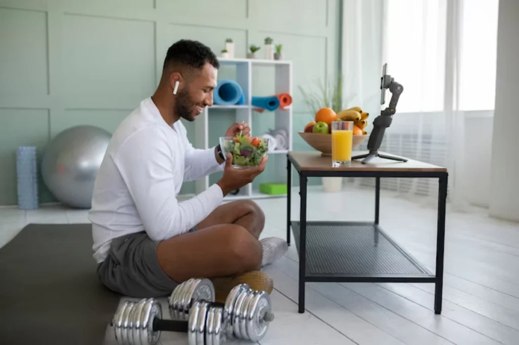 A person sits on a mat, enjoying a salad while a glass of juice and fresh fruit are on the table; dumbbells rest nearby.