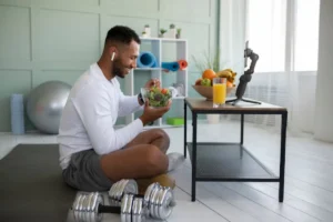 A person sits on a mat, enjoying a salad while a glass of juice and fresh fruit are on the table; dumbbells rest nearby.