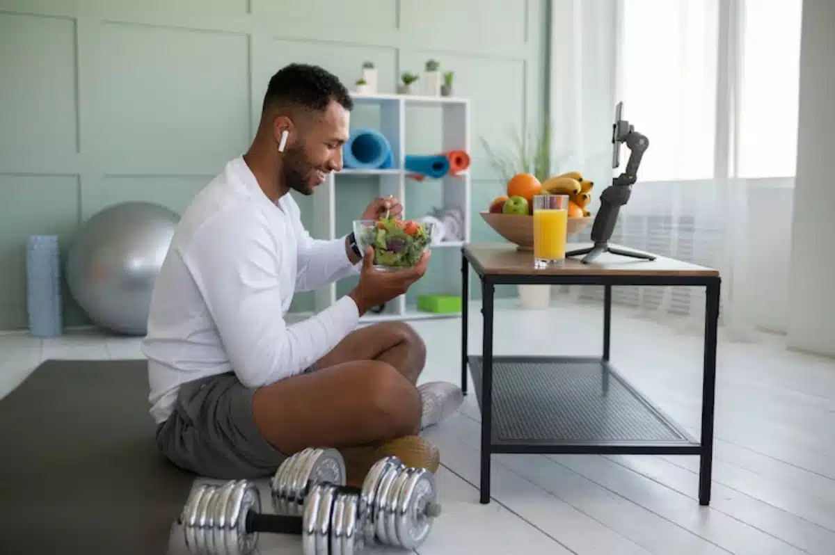 A person sits on a mat, enjoying a salad while a glass of juice and fresh fruit are on the table; dumbbells rest nearby.