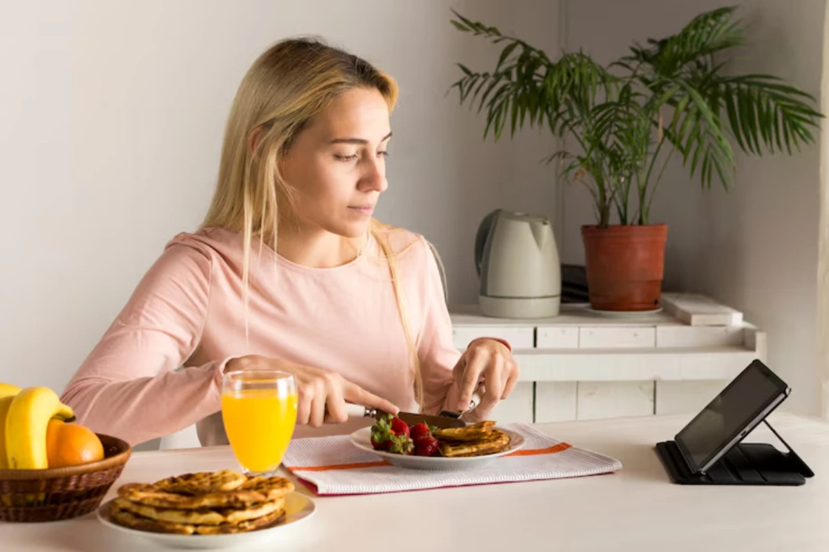 A person enjoys a breakfast of pancakes garnished with strawberries, alongside a glass of orange juice and a bowl of fruits.