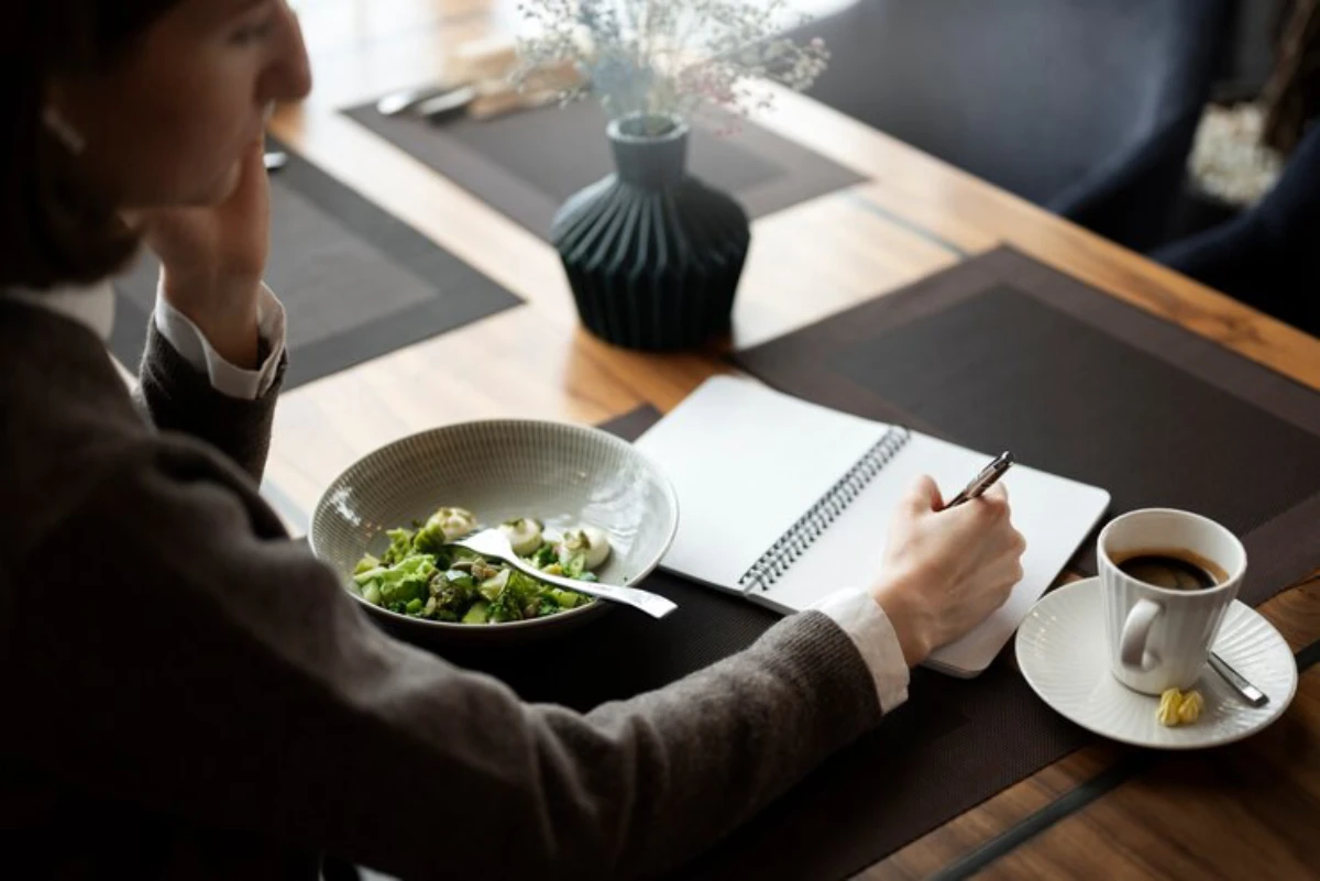 A person sits at a wooden table with a bowl of salad, a cup of coffee, and a notebook, writing with a pen. A vase adds a decorative touch.