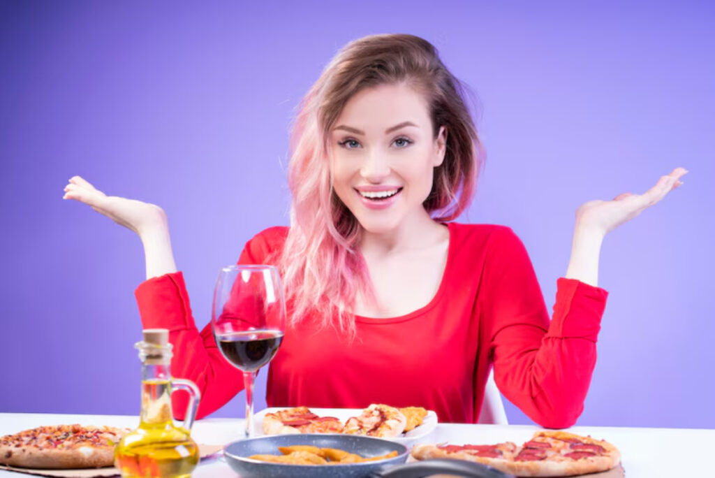 A person in a red top gestures with hands by a table filled with pizzas, snacks, and a glass of red wine, against a purple background.