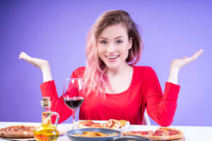 A person in a red top gestures with hands by a table filled with pizzas, snacks, and a glass of red wine, against a purple background.