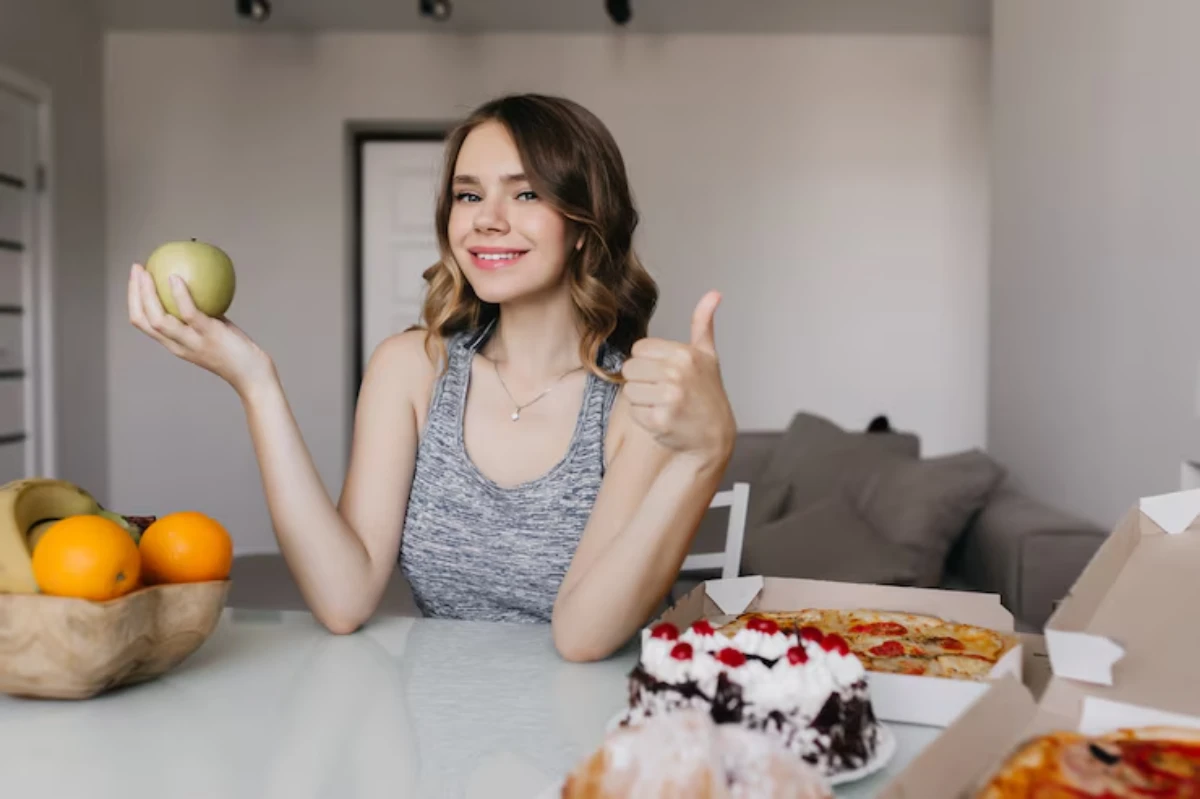 A woman holds a green apple in one hand and gives a thumbs up with the other, with a variety of food items on the table.