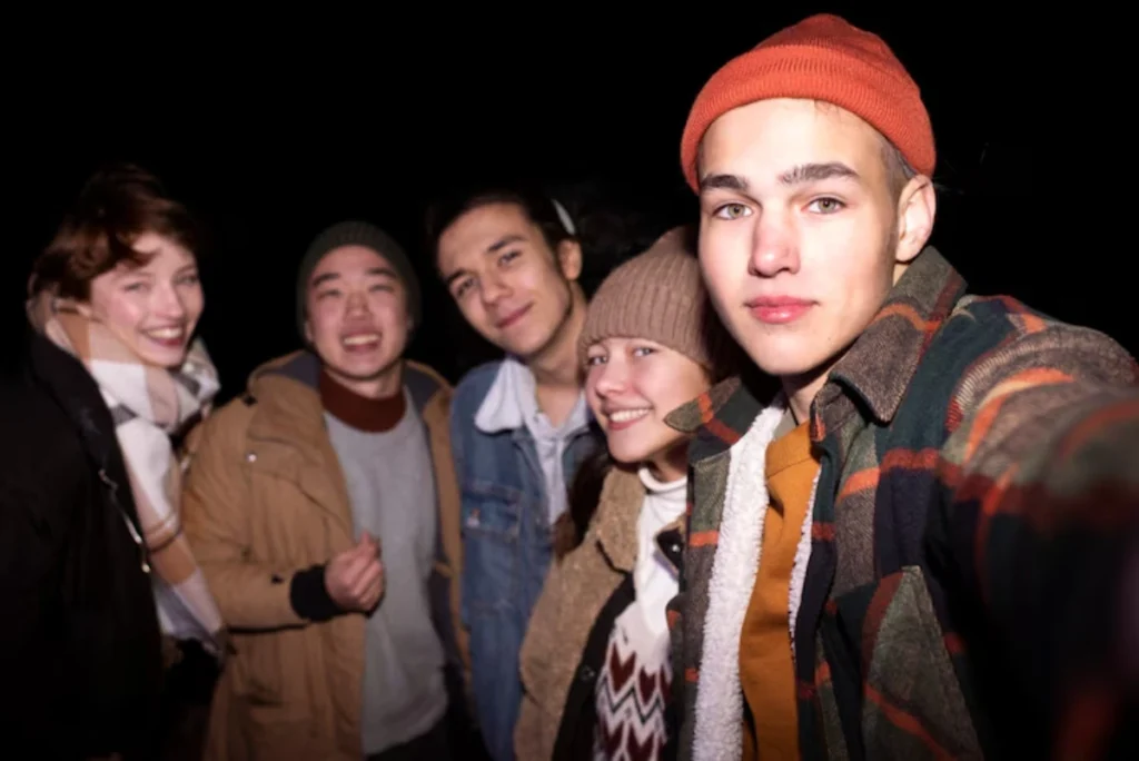 A group of five friends poses for a selfie at night, wearing casual, cozy outfits, surrounded by darkness.
