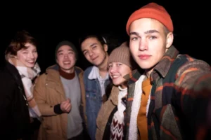 A group of five friends poses for a selfie at night, wearing casual, cozy outfits, surrounded by darkness.