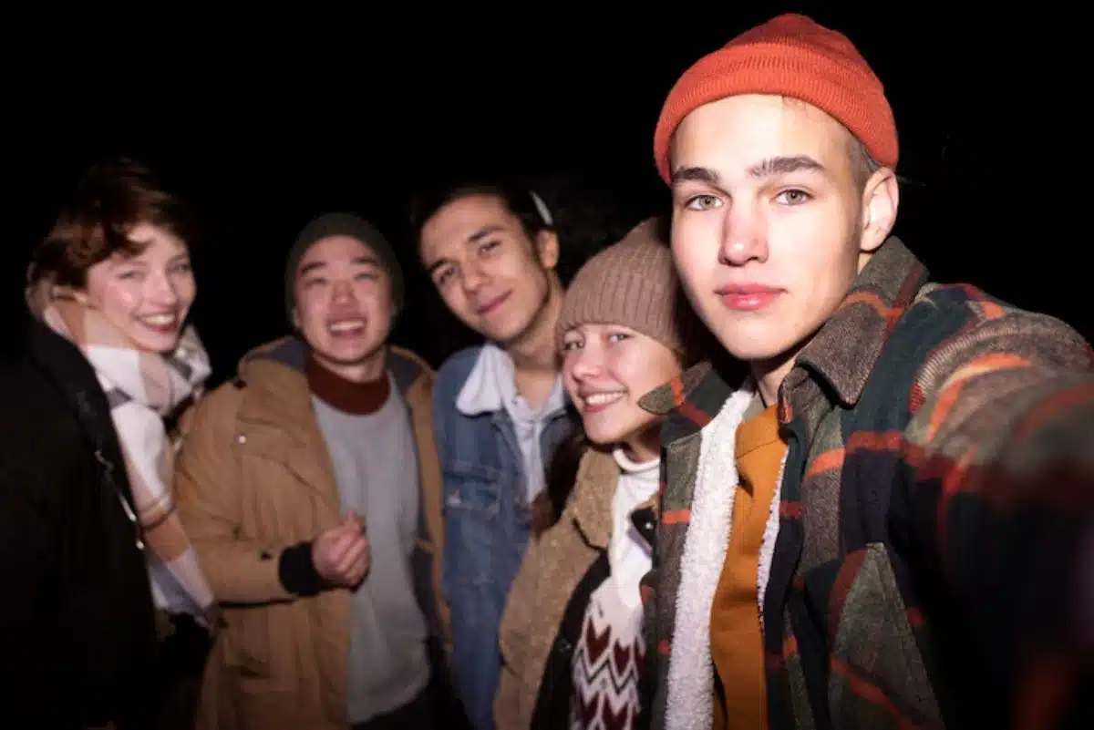 A group of five friends poses for a selfie at night, wearing casual, cozy outfits, surrounded by darkness.