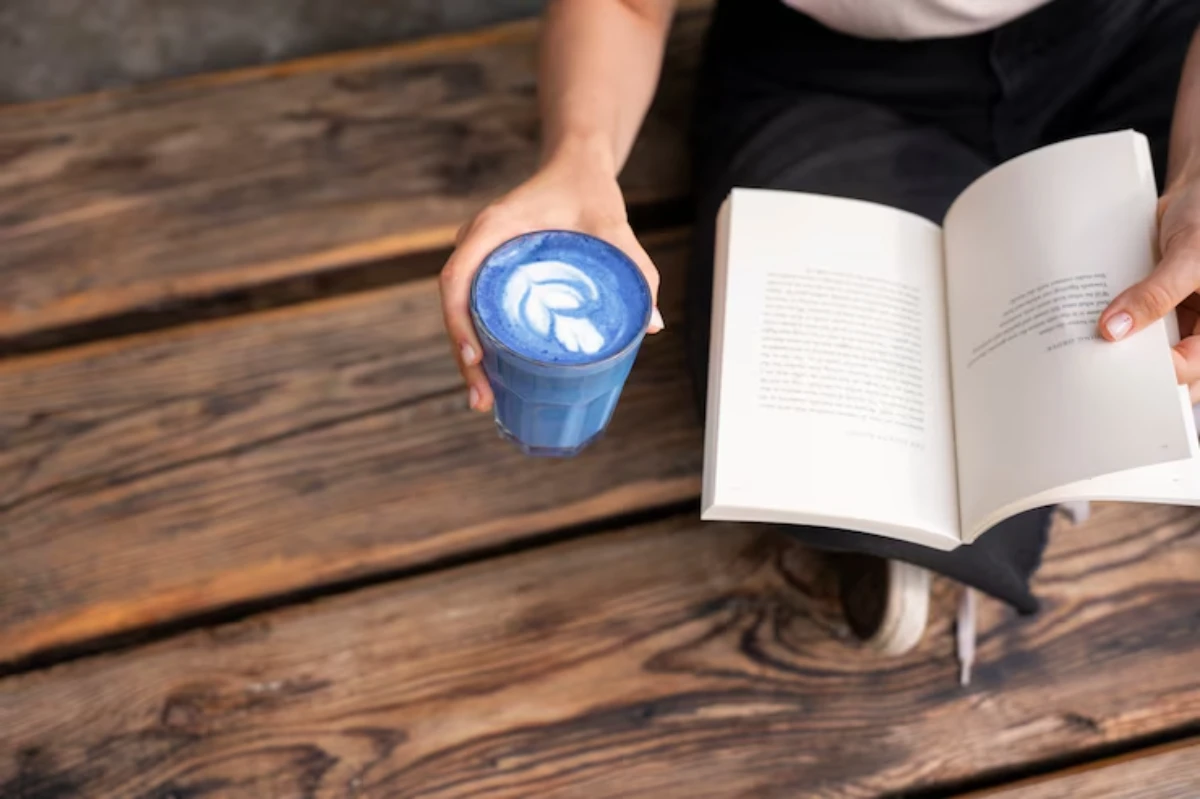 A person sitting on wooden planks holds a blue beverage with latte art in one hand and an open book in the other.