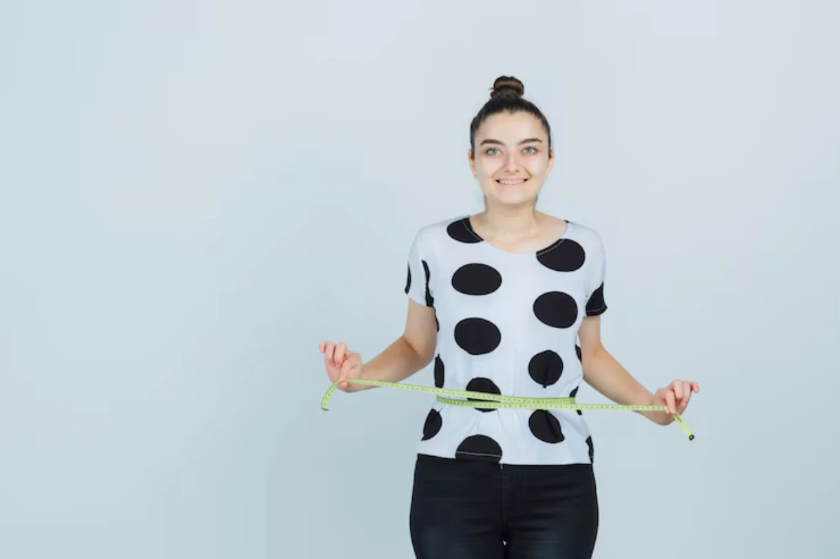 A woman in a polka dot shirt holds a measuring tape around her waist against a light blue background.