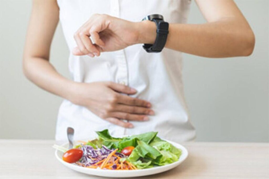 A person in a white shirt looks at their watch while holding their stomach, with a fresh salad on the table in front.