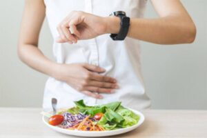A person in a white shirt looks at their watch while holding their stomach, with a fresh salad on the table in front.