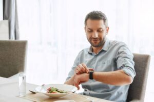 A person wearing a light blue shirt checks their watch while sitting at a table with a plate of salad and a glass of water.