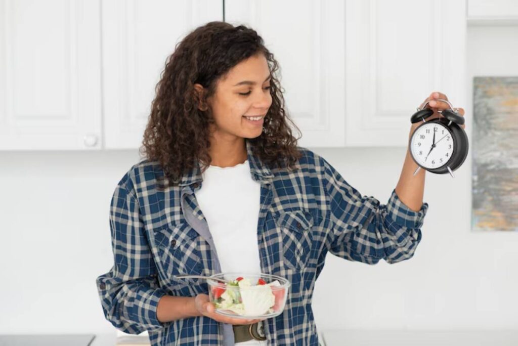 A person in a plaid shirt holds a clock in one hand and a bowl of salad in the other, standing in a modern kitchen.