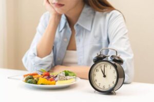 A woman sits at a table with a colorful plate of vegetables, looking contemplative next to a black alarm clock.